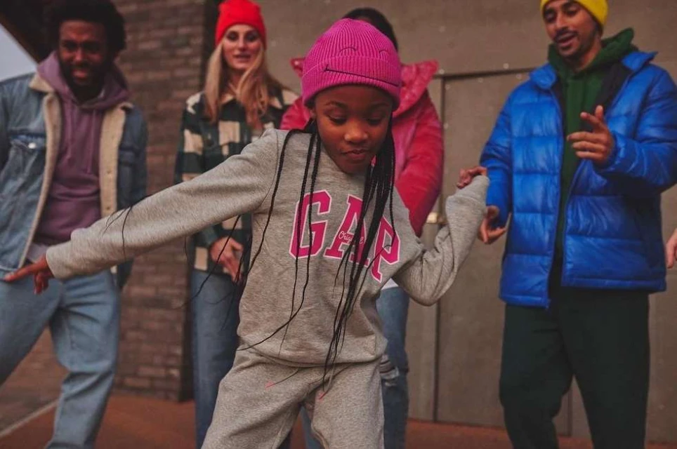 A young girl wearing a gray GAP tracksuit dances in front of a diverse group of people in colorful hats and casual clothing outdoors.