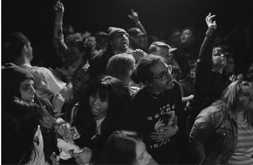 Crowd of people at a concert, with several individuals raising their hands in the air, captured in a high-contrast black and white photo.