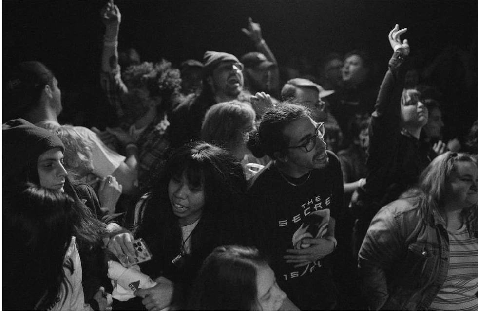 Crowd of people at a concert, with several individuals raising their hands in the air, captured in a high-contrast black and white photo.