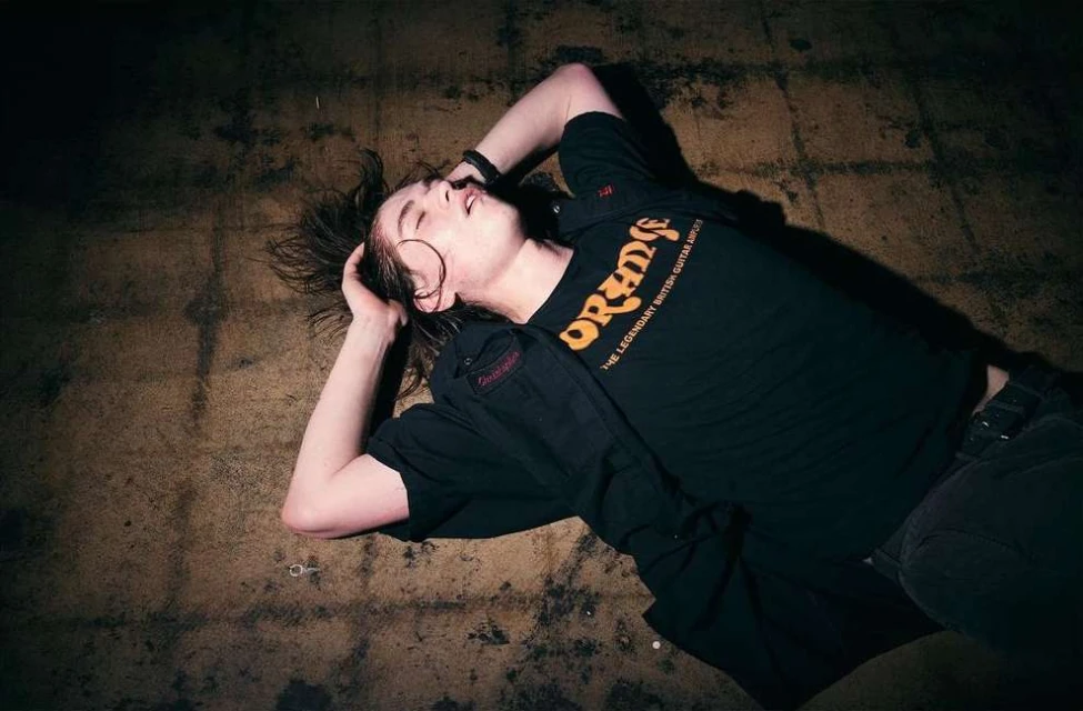 A young adult male lies on the floor with hands behind his head, wearing a black t-shirt with an orange logo and dark jeans.