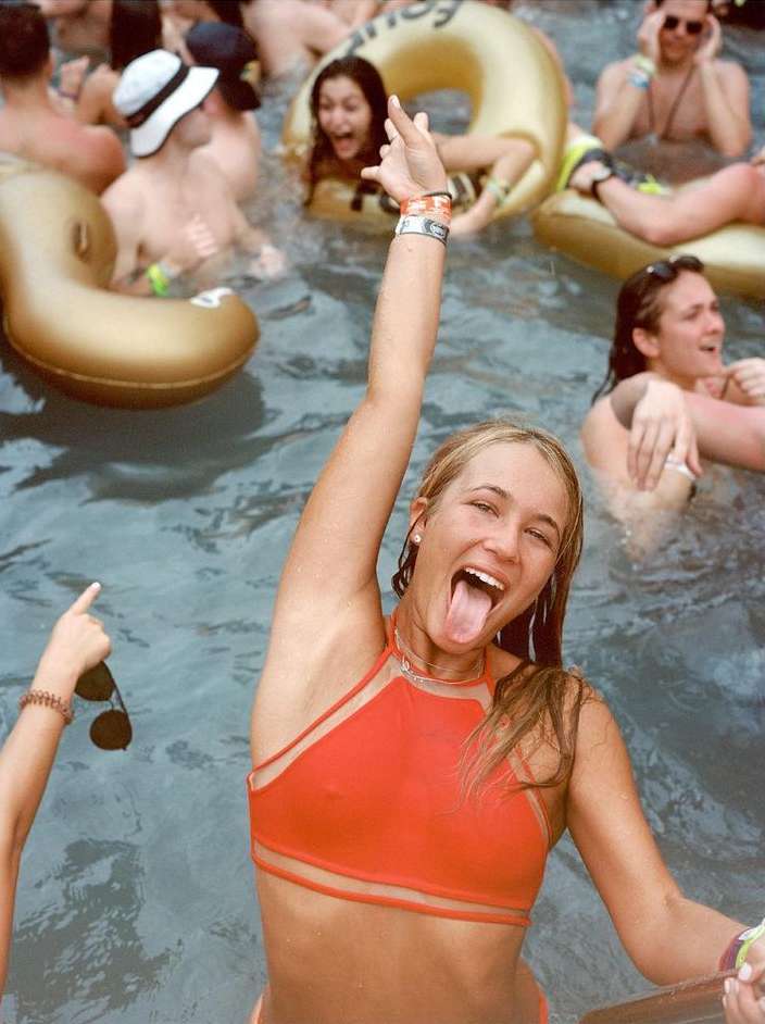 Young woman in orange bikini top in a pool party with friends, sticking tongue out and raising hand in celebratory gesture.