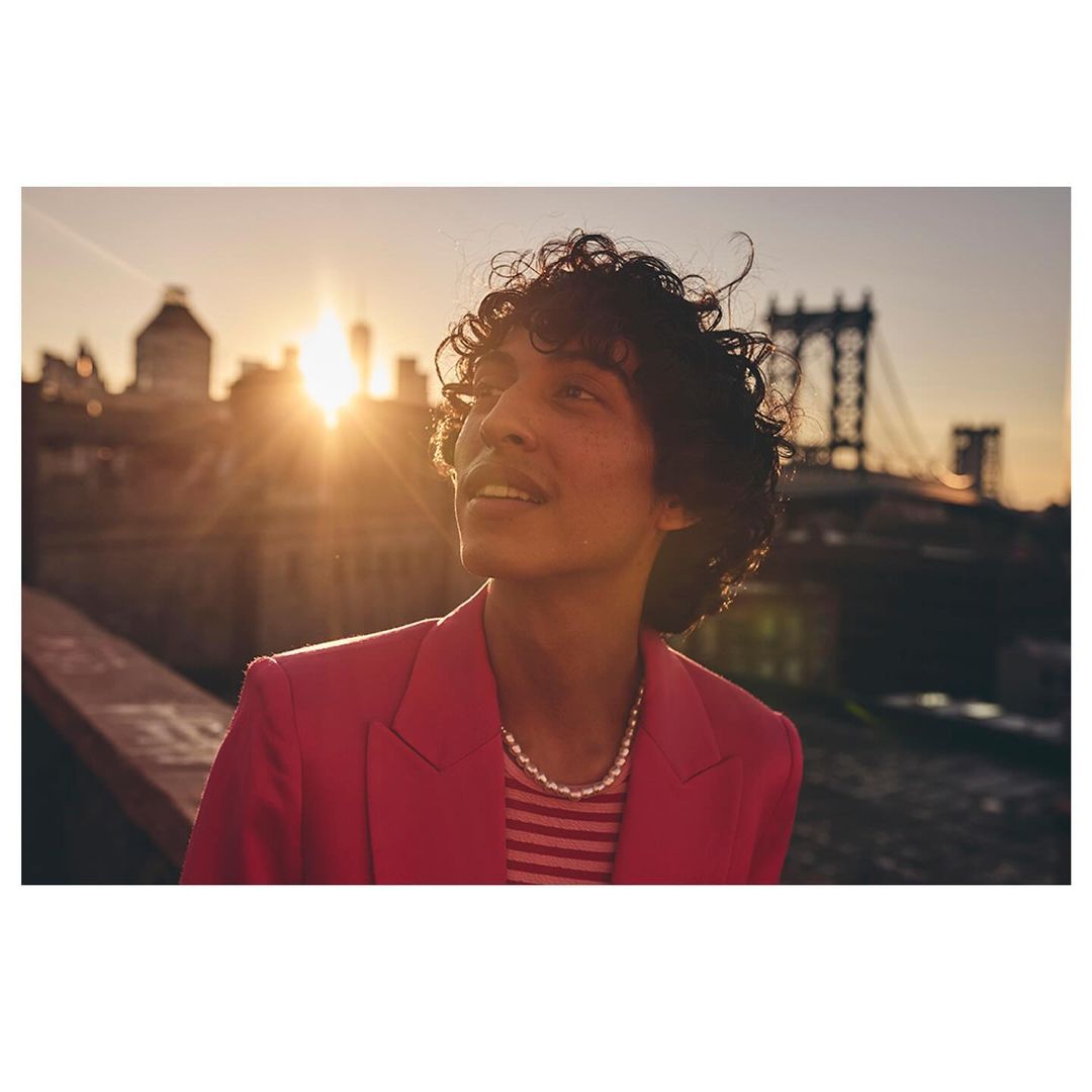 Portrait of a young adult male with curly hair in a pink jacket and pearl necklace looking upward, with a city skyline at sunset.