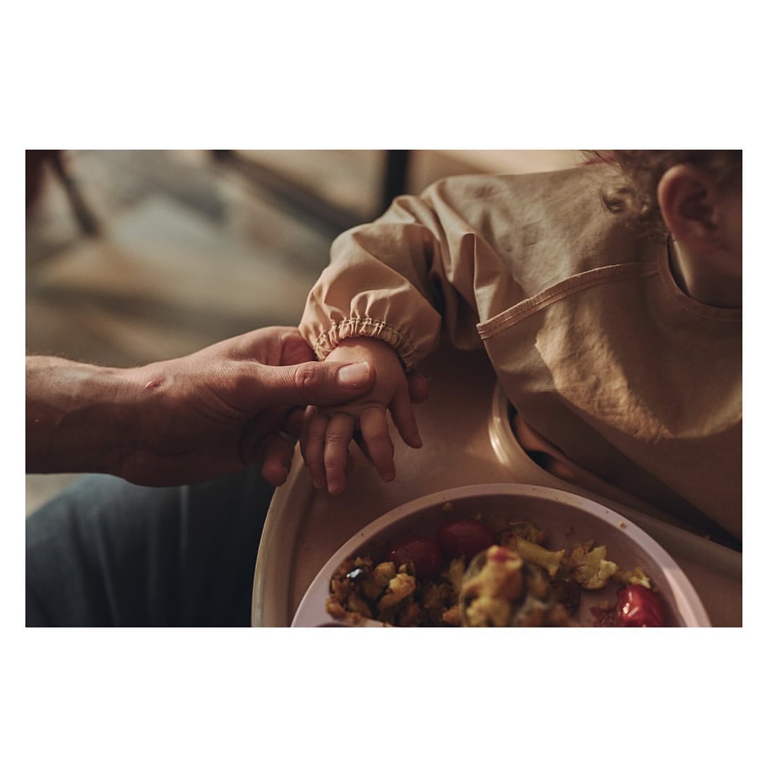 Adult's hand holding a baby's hand as the child sits in a high chair with a plate of food with tomatoes and mixed vegetables.