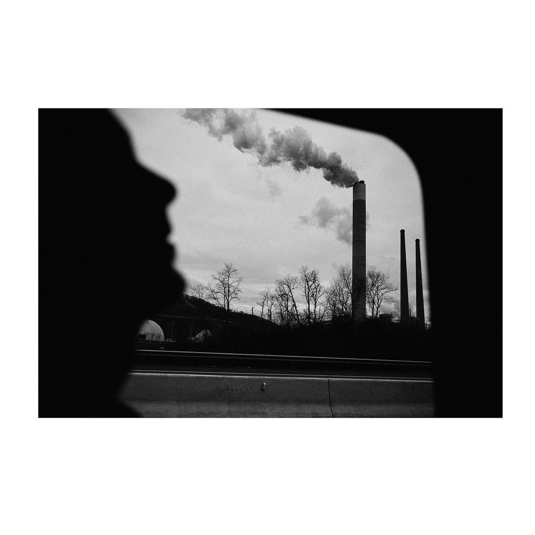 Silhouette of a person gazing out a car window at industrial smokestacks emitting smoke against a cloudy sky, creating a monochrome effect.