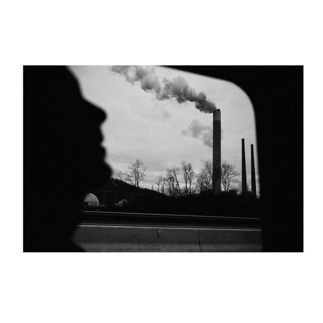 Silhouette of a person gazing out a car window at industrial smokestacks emitting smoke against a cloudy sky, creating a monochrome effect.