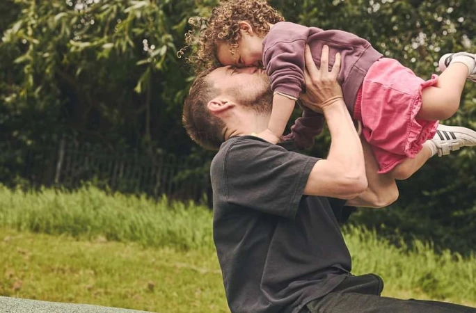 Man lifting a young child up in the air outdoors with green grass and trees in the background