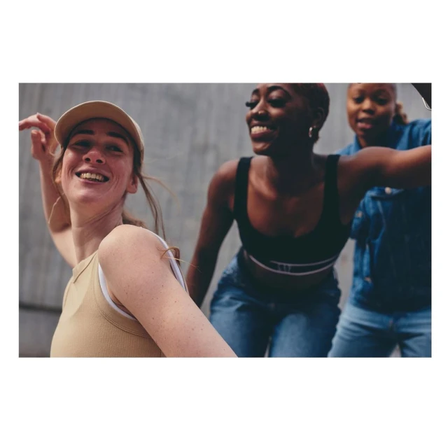 Three young women in casual clothing smile and pose against a textured grey wall, bathed in soft, natural light, expressing youthful energy.