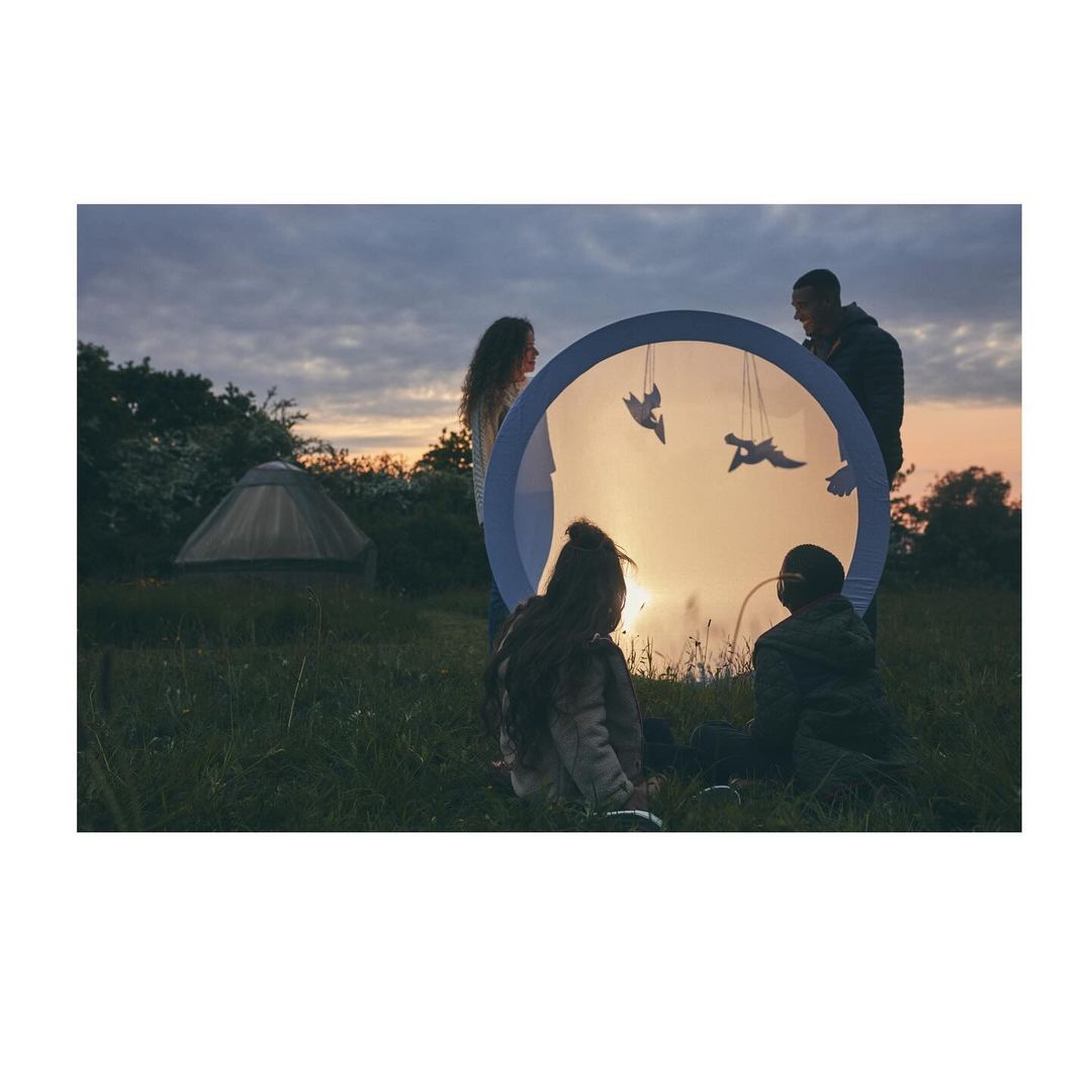 Family gathers in a grassy field at dusk, enjoying an illuminated, transparent sphere with bird silhouettes.  A yurt is visible.