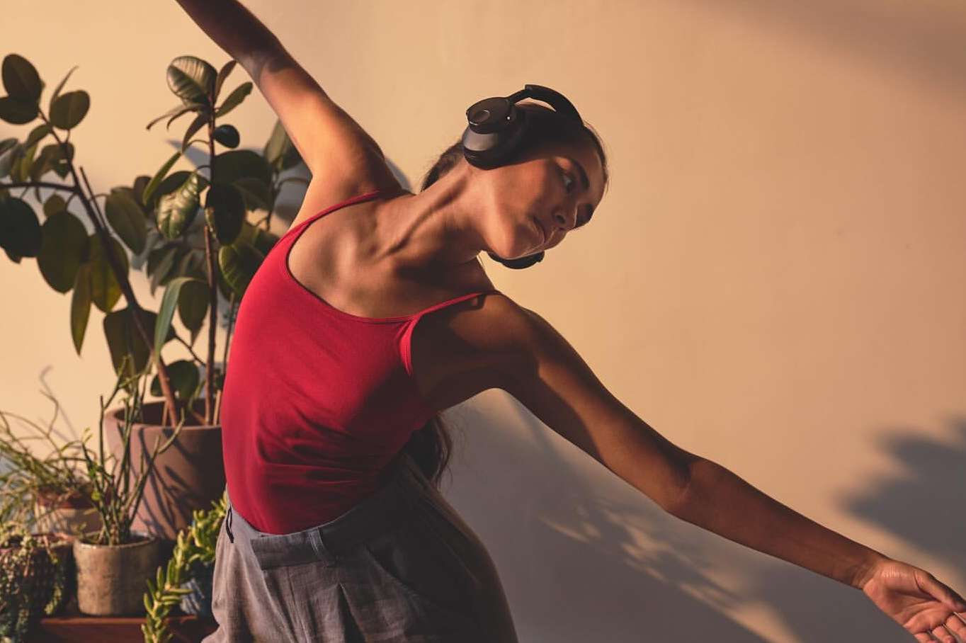 Woman in red top and grey pants wears headphones in a dance pose next to indoor plants against a peach-toned wall in a studio setting.