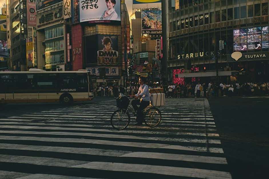 Man cycling across a crosswalk in a bustling urban area, featuring a bus, crowds, billboards, and Starbucks in the background.
