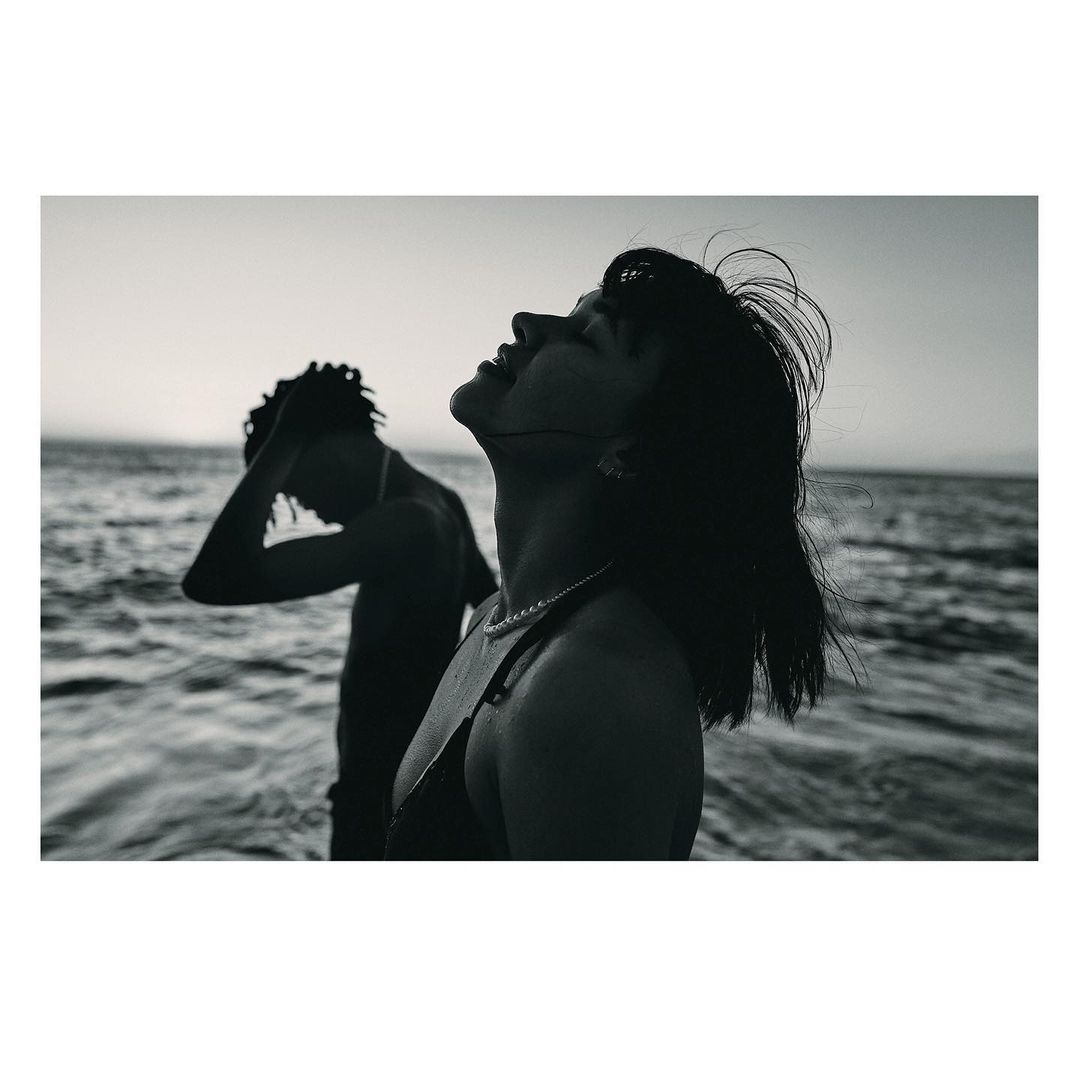 Monochrome portrait of a woman with dark hair tilting her head back, with a man standing in the ocean surf behind her.