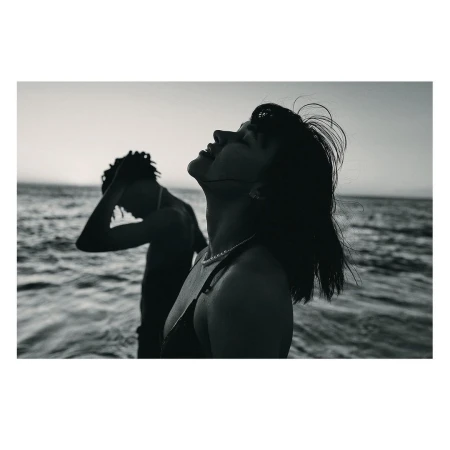 Monochrome portrait of a woman with dark hair tilting her head back, with a man standing in the ocean surf behind her.