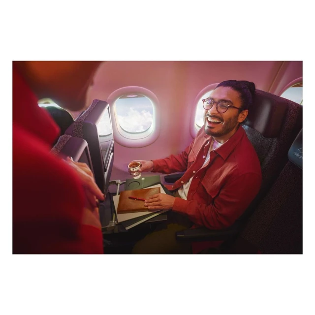 A man in a plane smiles while holding a drink, with another person visible on the left in an airline interior. 