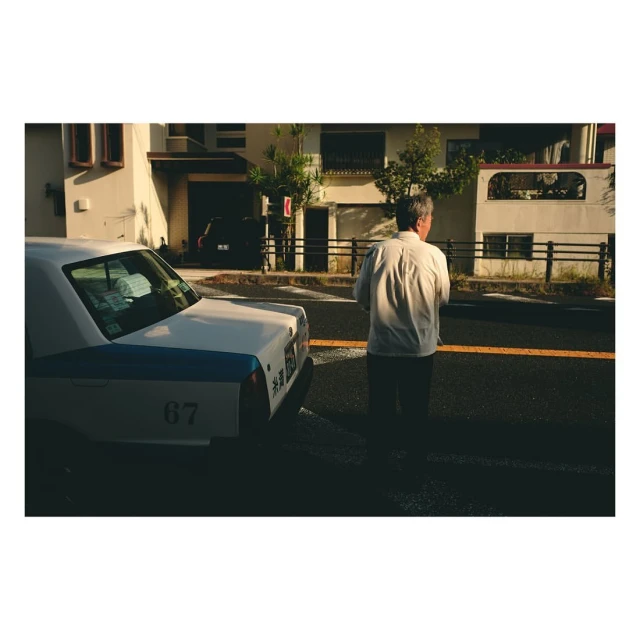 Mature man in a button-down shirt standing on a street with a white taxi and buildings in the background during daylight hours.