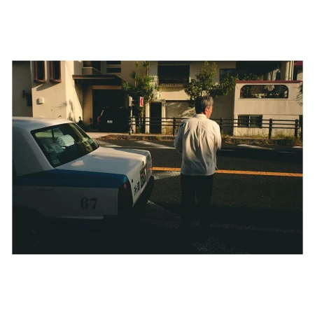 Mature man in a button-down shirt standing on a street with a white taxi and buildings in the background during daylight hours.