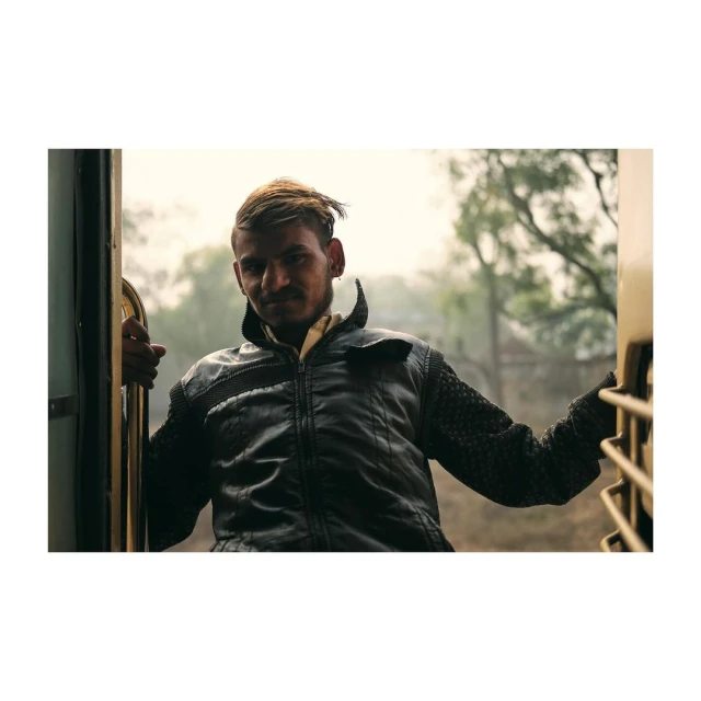 Portrait of a man leaning out of a train doorway, wearing a black jacket with textured sleeves, set against a soft, outdoor background.