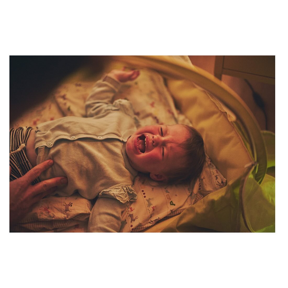 A baby lies on a patterned blanket with a distressed facial expression while an adult's hand rests near the infant's lower body.