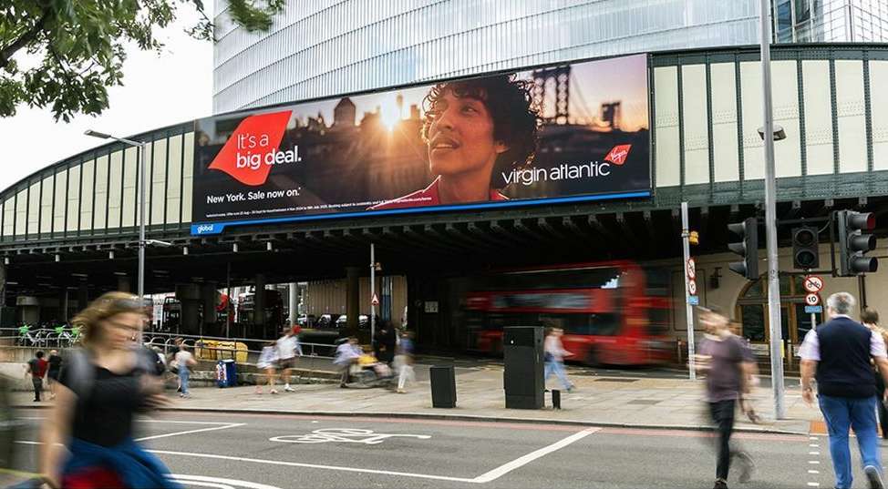 Virgin Atlantic billboard features a man in a city setting with blurred pedestrians and a red double-decker bus passing by.