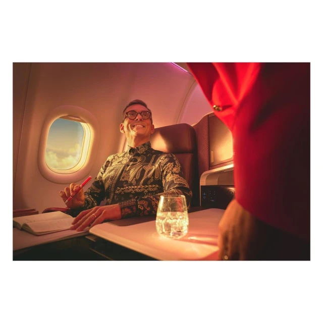 Man smiles inside airplane, seated by window. He wears glasses and patterned shirt, with glass of water and book on the table.