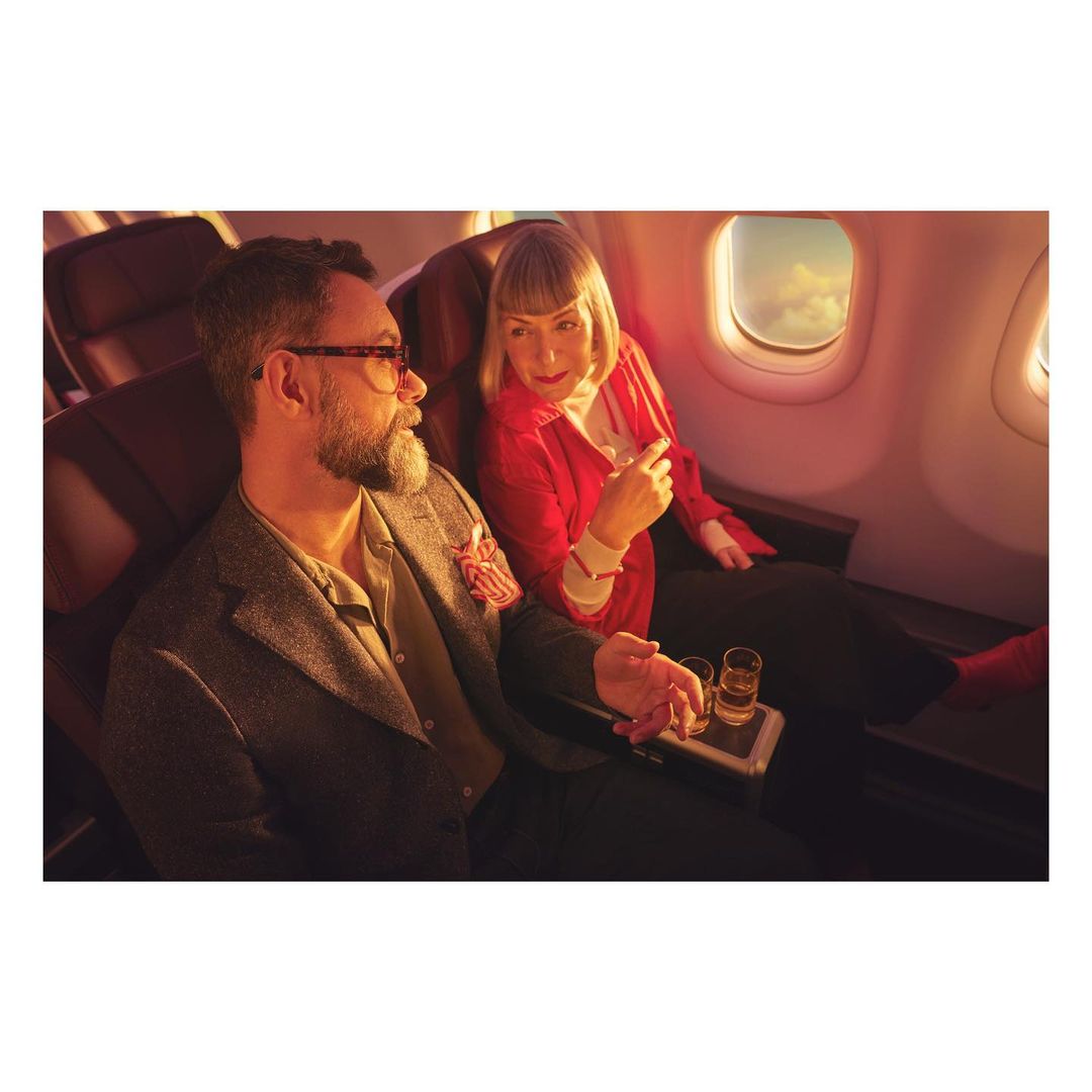 Adult male and female seated on an airplane with drinks. Man in glasses and grey jacket looks at woman wearing red jacket and black pants.