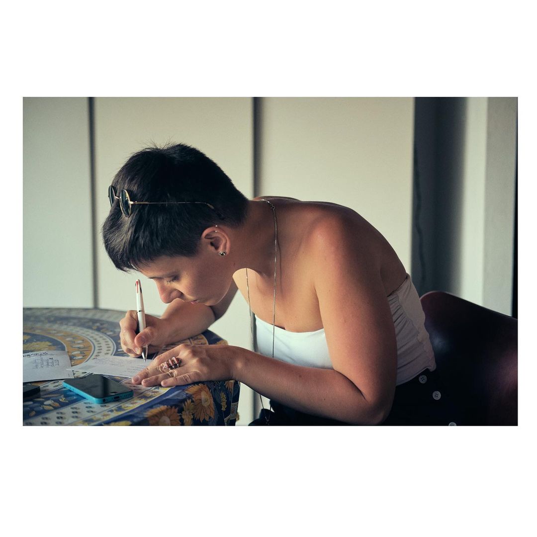 A woman with short dark hair is seated at a round table writing on a piece of paper, with a smartphone and some papers nearby.