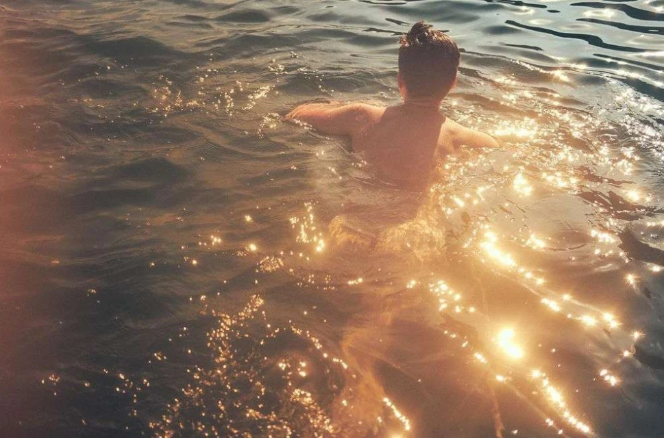 Back view of person swimming in water with sunlight creating sparkling reflections, outdoors in nature during daytime.
