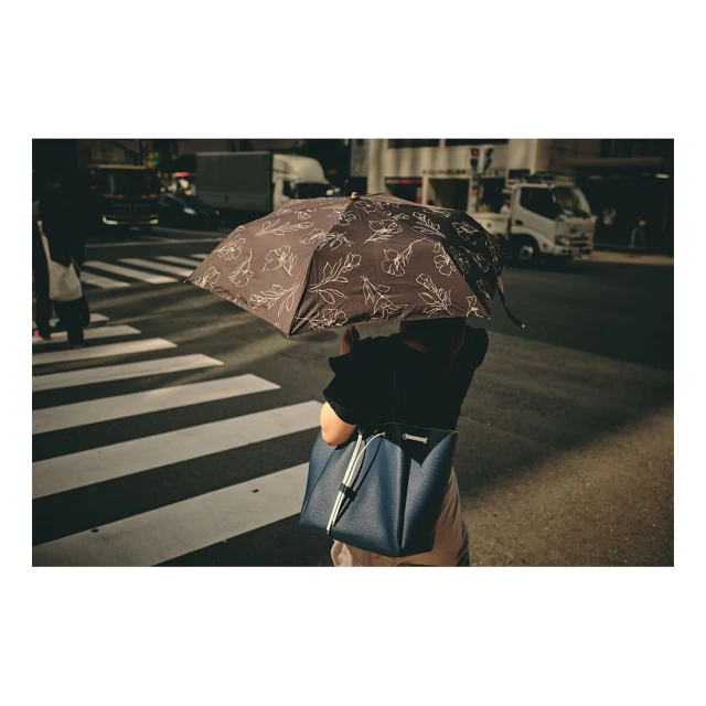 Person walking on crosswalk with a brown floral-print umbrella and blue shoulder bag in an urban environment with city traffic.