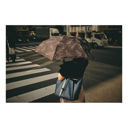 Person walking on crosswalk with a brown floral-print umbrella and blue shoulder bag in an urban environment with city traffic.