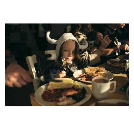 Child in costume eating breakfast at a table with family, featuring plates of food, coffee, and natural window lighting.