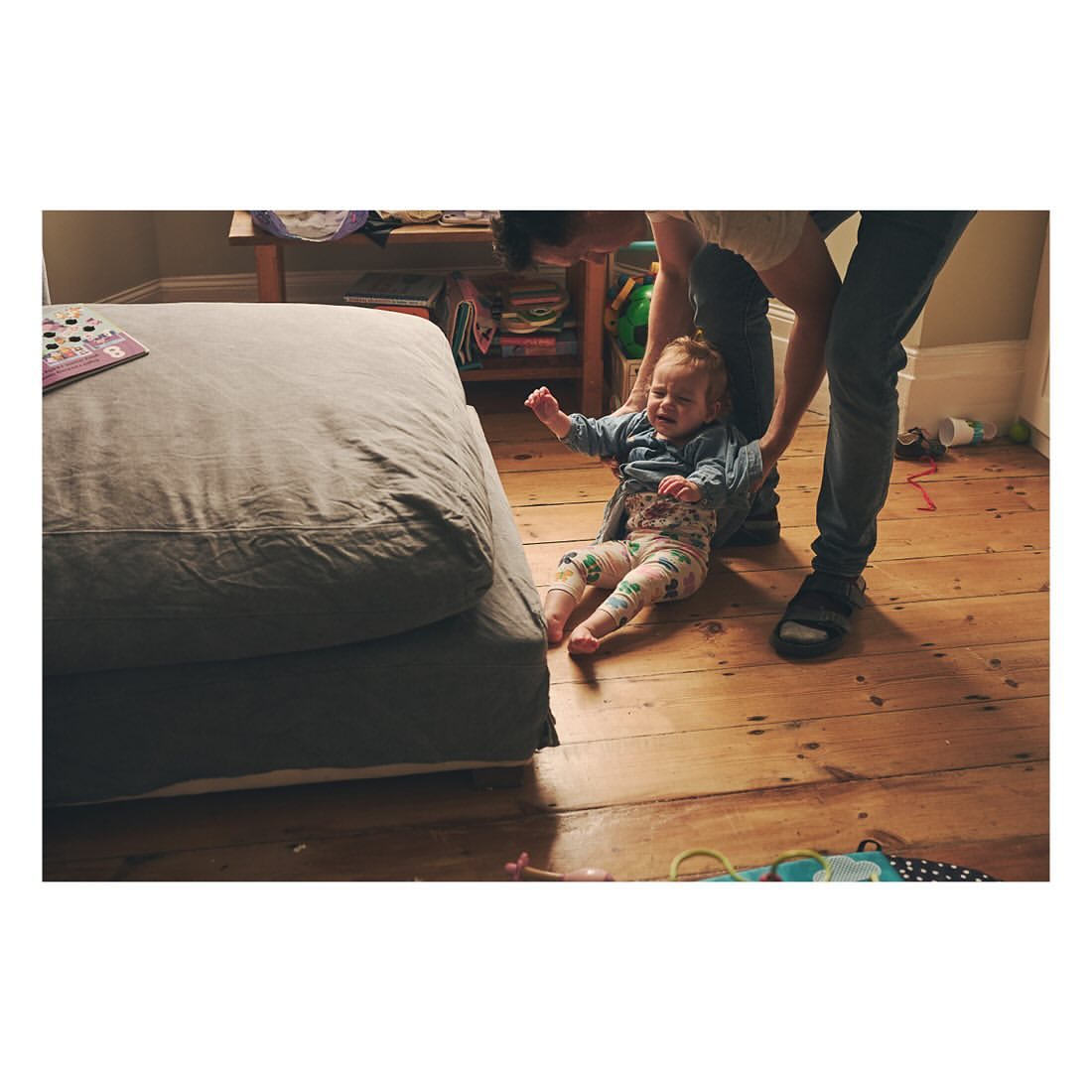 A toddler cries while being held by an adult in an indoor room with a hardwood floor and a gray mattress on the floor.
