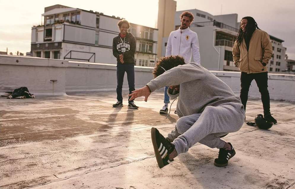 A group of four young adults stands on a flat rooftop on a sunny day. One man in a grey sweat suit is breakdancing in the foreground.