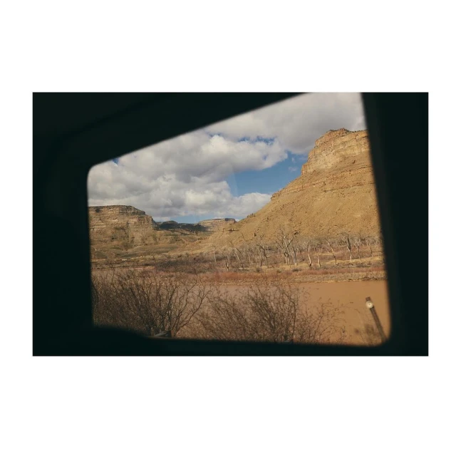 Scenic view of a tan cliffside under a blue and cloudy sky, as seen through a dark square window of a vehicle.