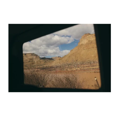 Scenic view of a tan cliffside under a blue and cloudy sky, as seen through a dark square window of a vehicle.
