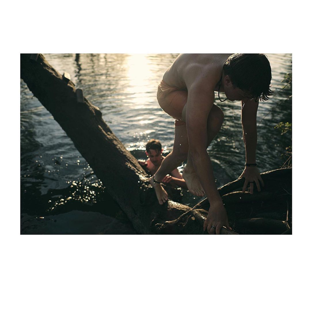 Two young men on log in lake under bright sun. One man climbs onto the log from the water, another swims nearby in daytime. 