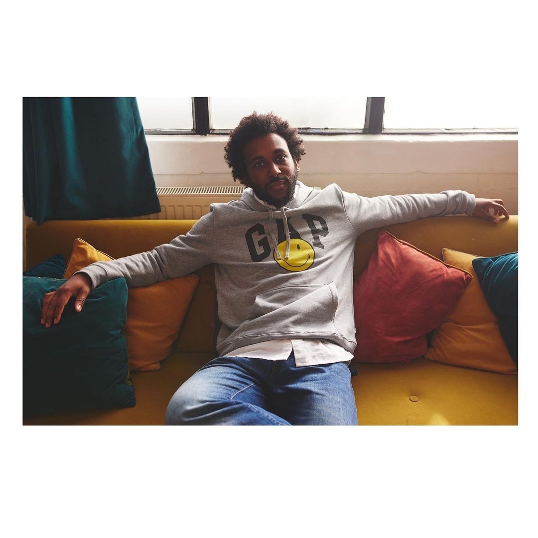 Adult male sitting on a yellow couch wearing a gray GAP smiley-face hoodie, jeans, and white shirt. He is posing indoors.