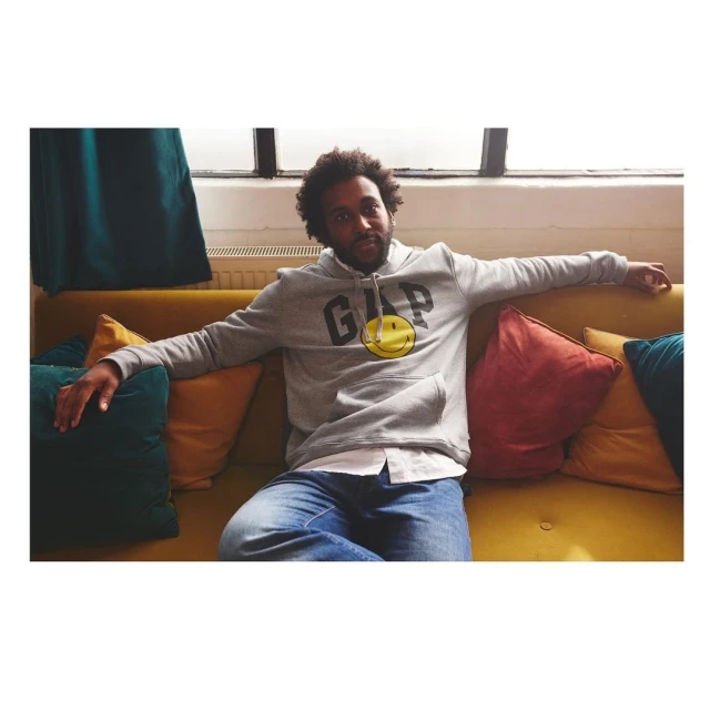 Adult male sitting on a yellow couch wearing a gray GAP smiley-face hoodie, jeans, and white shirt. He is posing indoors.