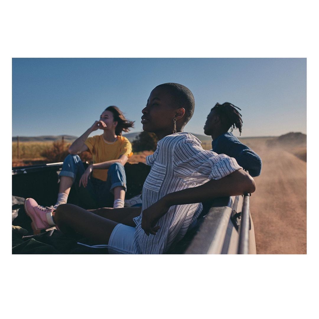 Three young adults in the back of an open vehicle driving on a rural dirt road under a clear blue sky, captured in natural daylight.