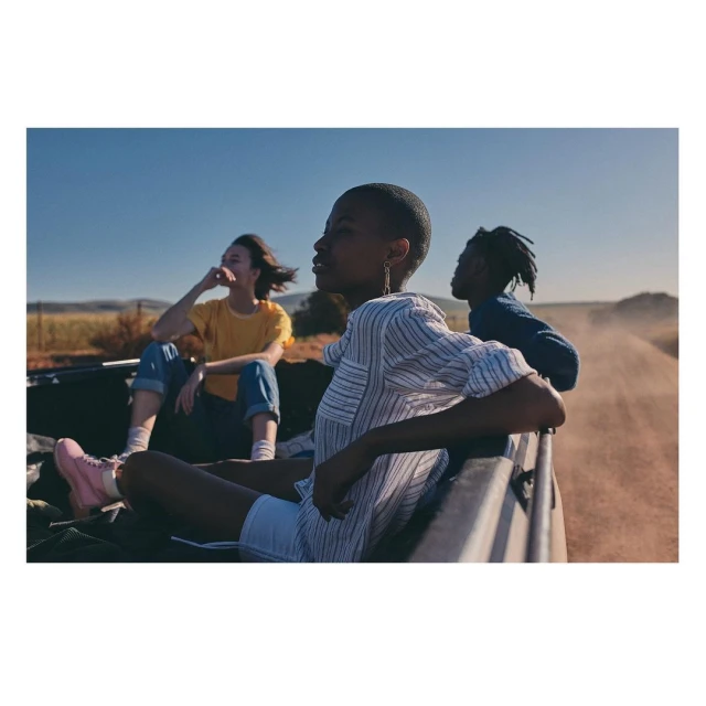 Three young adults in the back of an open vehicle driving on a rural dirt road under a clear blue sky, captured in natural daylight.
