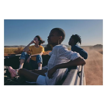 Three young adults in the back of an open vehicle driving on a rural dirt road under a clear blue sky, captured in natural daylight.
