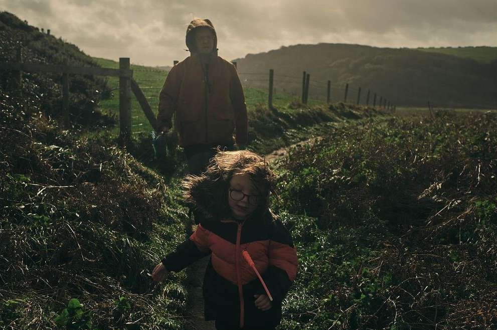 A child and adult walk a grassy path near a fence and hillside; the child wears glasses and a fur-trimmed coat, a cloudy day.