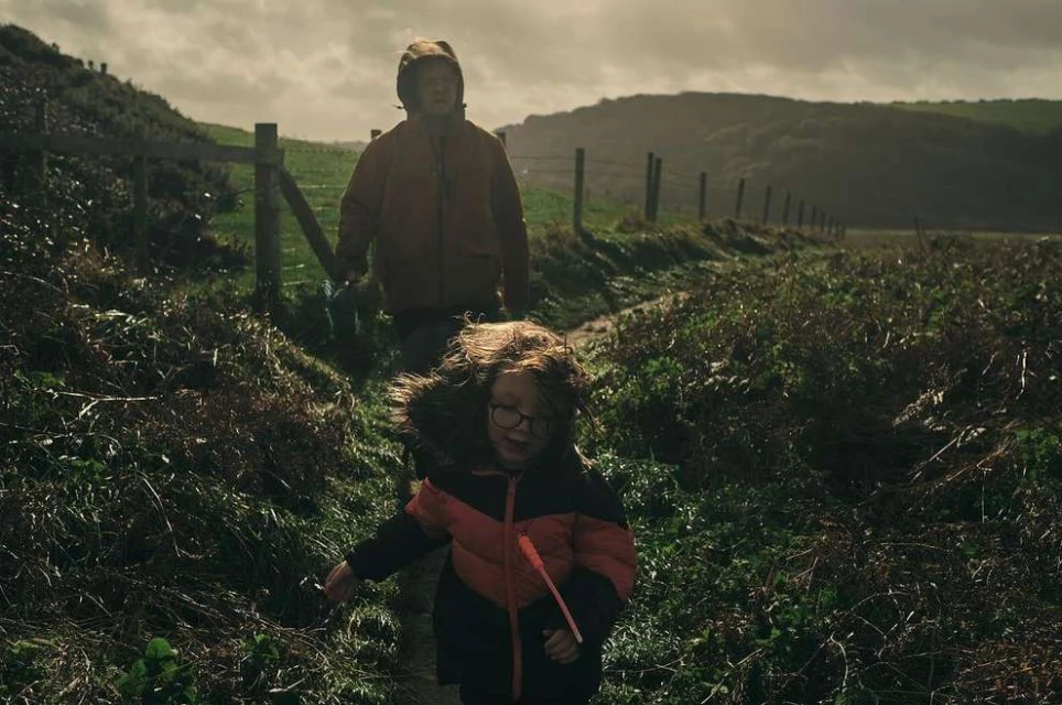 A child and adult walk a grassy path near a fence and hillside; the child wears glasses and a fur-trimmed coat, a cloudy day.