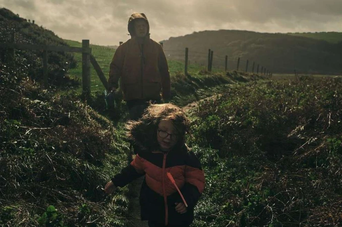 A child and adult walk a grassy path near a fence and hillside; the child wears glasses and a fur-trimmed coat, a cloudy day.