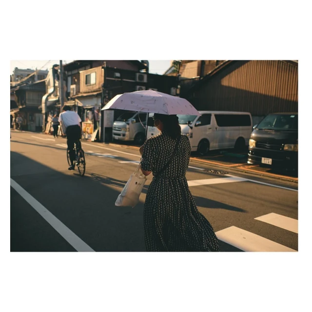 Woman crosses street under umbrella as cyclist rides by in urban setting with buildings and parked cars in warm afternoon light.