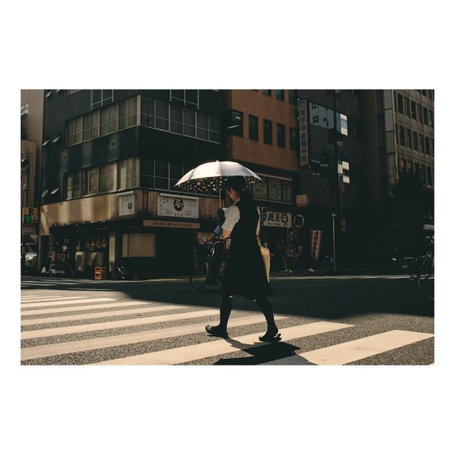 Woman with umbrella walking across crosswalk in Tokyo with buildings, street signs, and bicycles visible in the urban setting.