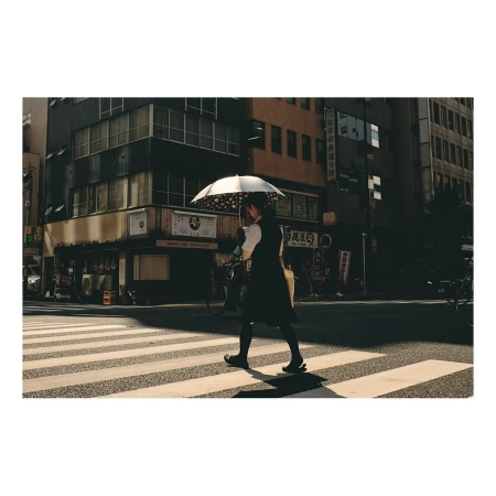 Woman with umbrella walking across crosswalk in Tokyo with buildings, street signs, and bicycles visible in the urban setting.