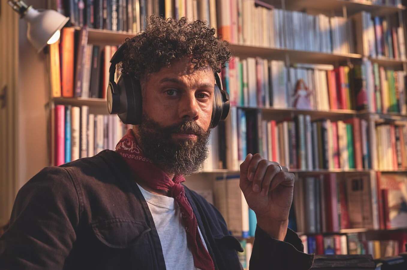 Portrait of an adult male with a beard, wearing headphones and a neck scarf, posing indoors with a bookshelf full of books in the background.