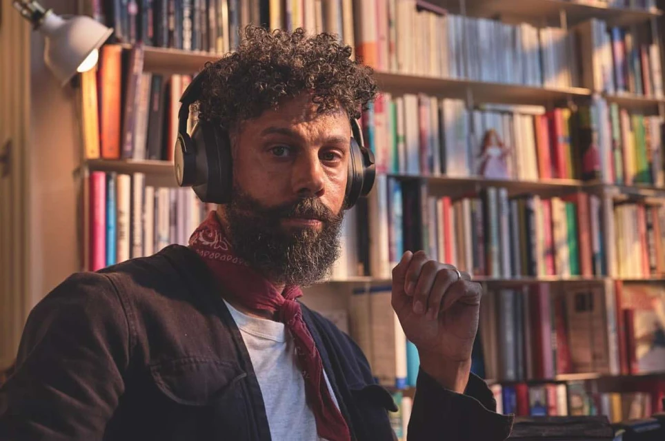 Portrait of an adult male with a beard, wearing headphones and a neck scarf, posing indoors with a bookshelf full of books in the background.