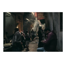 Man in purple jacket seated in a narrow street filled with figures, smoke, and storefronts, captured by Will Hartley.