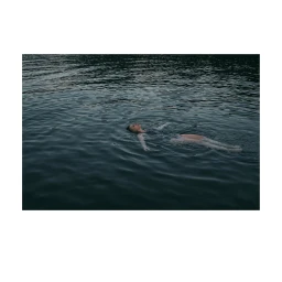 A person floating on their back in dark water with arms outstretched under an overcast sky.