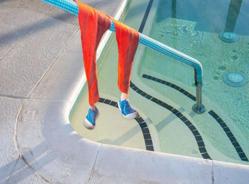 Orange pants hang on pool railing with blue shoes just above water, showcasing unique poolside fashion and refreshing summer vibes.
